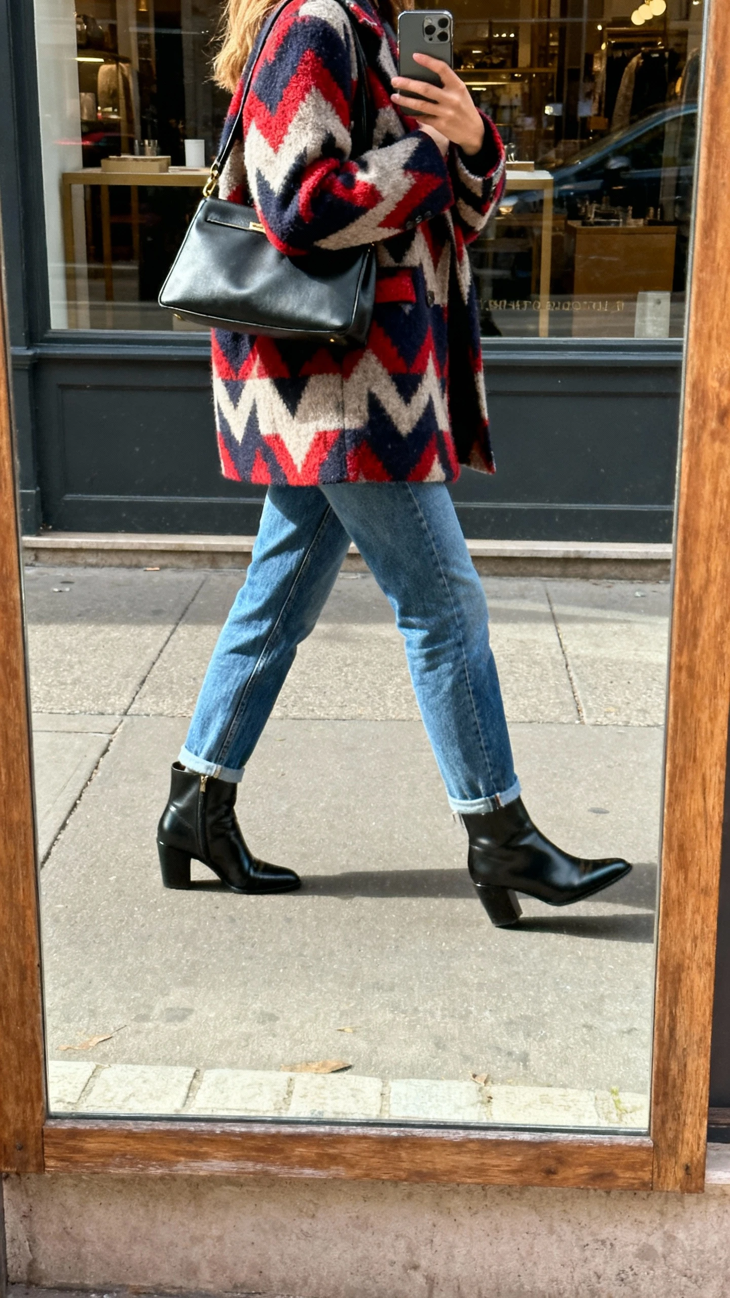 iPhone street-level mirror selfie (storefront reflection) of a woman wearing mom jeans, a bold statement coat, sleek ankle boots, structured handbag, face not visible, back/side view walking pose, natural daylight, iPhone photo quality