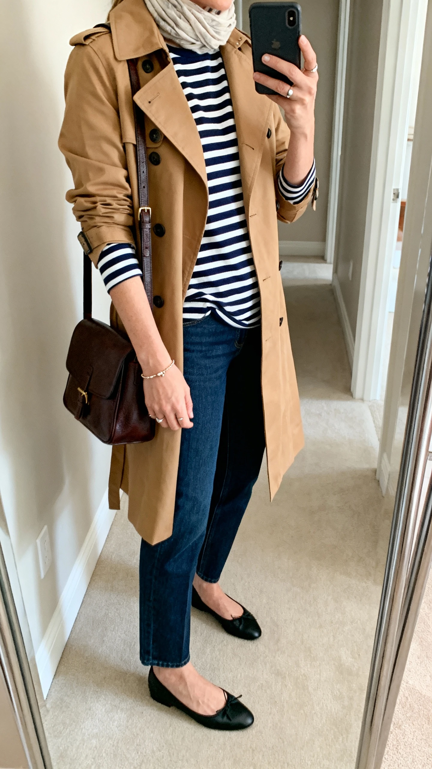 Side-angle iPhone mirror selfie of a woman wearing a camel trench coat over a navy-and-white Breton stripe long-sleeve tee, ankle-length straight-leg jeans, black ballet flats, structured leather shoulder bag, delicate bracelet, simple studs, and a lightweight scarf; face not visible, cropped at shoulders, hallway full-length mirror, soft morning light, iPhone photo quality.