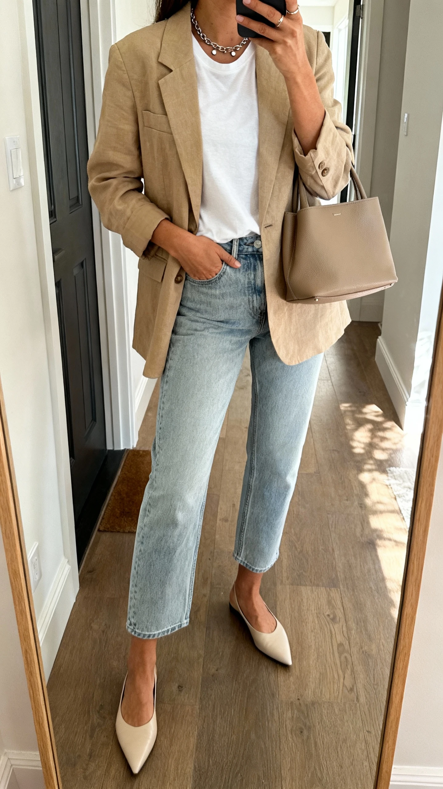 Side-angle iPhone mirror selfie of a woman wearing an unstructured beige blazer over a high-quality white tee, straight-leg light-wash cropped jeans, pointed flats, minimalist leather tote, simple studs and delicate chain necklace, face not visible, cropped at shoulders, entryway full-length mirror, natural afternoon light, iPhone photo quality.